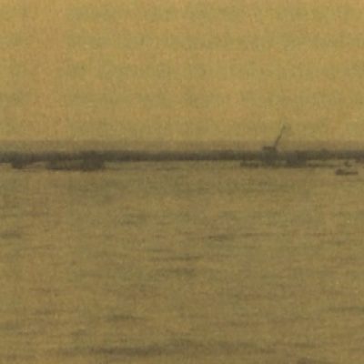 Historic photograph of a dredging or sediment-placement operation at Fowl River, showing a long pipeline or barge system extending across open water between work vessels and a pile of material.
