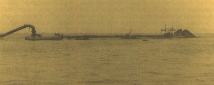 Historic photograph of a dredging or sediment-placement operation at Fowl River, showing a long pipeline or barge system extending across open water between work vessels and a pile of material.