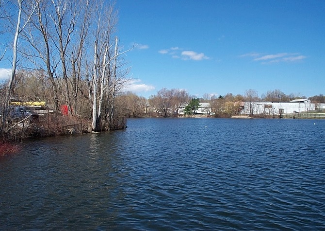 View across Silver Lake, with open blue water bordered by leafless trees and shoreline vegetation. Residential and industrial buildings are visible along the opposite shore beneath a clear blue sky.