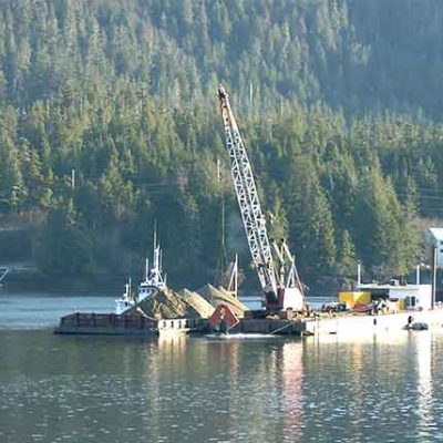 Marine construction operation on calm water, with a crane mounted on a barge handling material or equipment. Additional vessels and an industrial facility sit near the shoreline, backed by a densely forested hillside.