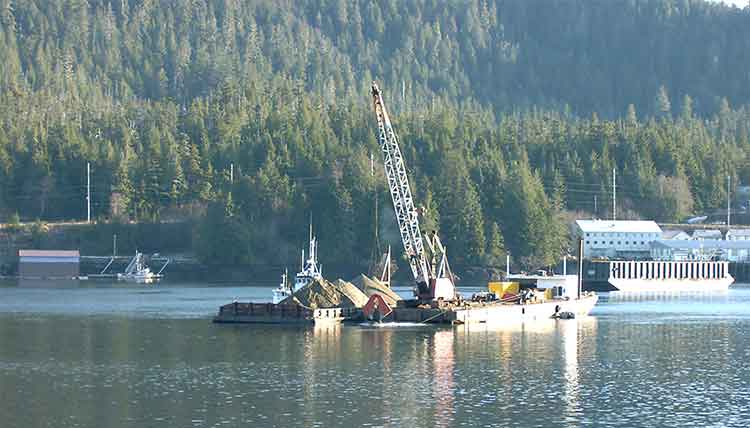 Marine construction operation on calm water, with a crane mounted on a barge handling material or equipment. Additional vessels and an industrial facility sit near the shoreline, backed by a densely forested hillside.