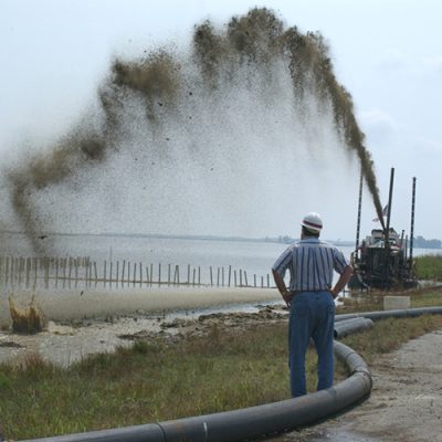 Sediment slurry is sprayed in a high arc from pumping equipment into a shallow wetland at Blackwater, while two workers in hard hats observe beside a large pipeline.