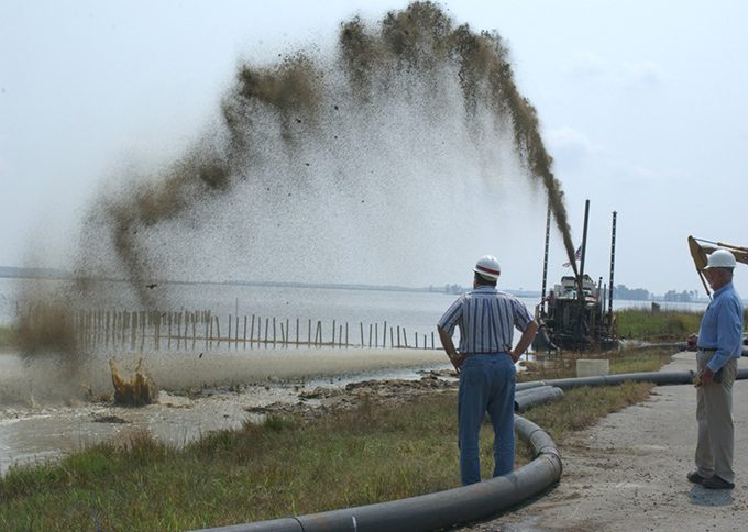Sediment slurry is sprayed in a high arc from pumping equipment into a shallow wetland at Blackwater, while two workers in hard hats observe beside a large pipeline.