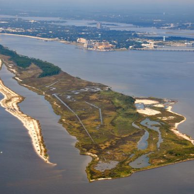 Aerial view of Deer Island showing marsh habitat, tidal channels, sandy shoreline, and surrounding coastal waters, with developed shoreline in the distance.