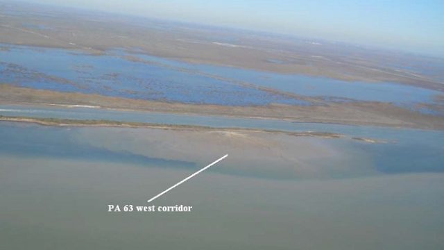Aerial view of the PA 63 West Corridor, showing a narrow constructed shoreline beside a navigation channel, with broad expanses of shallow water and coastal marsh beyond. An arrow identifies the west corridor.