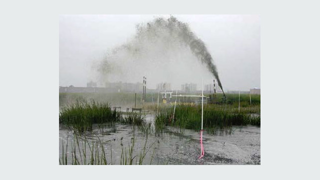 Sediment slurry is sprayed in a high arc across a shallow wetland at Pepper Creek, with marsh vegetation and monitoring stakes visible around the placement area.