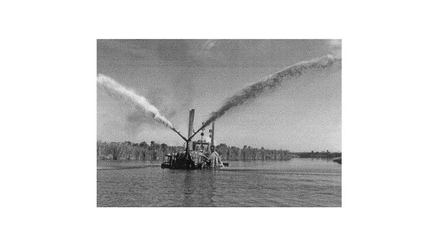 Black-and-white photograph of a floating sediment-placement vessel spraying two large arcs of slurry in opposite directions across a shallow wetland restoration area.