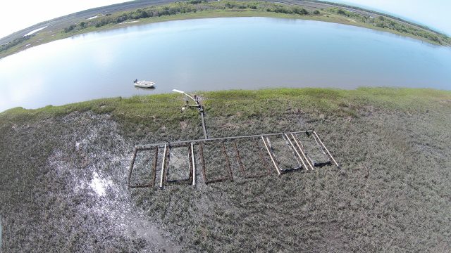 Aerial view of experimental plots arranged in rows across a tidal marsh beside a broad waterway, with a small boat near shore and monitoring equipment positioned among the plots.