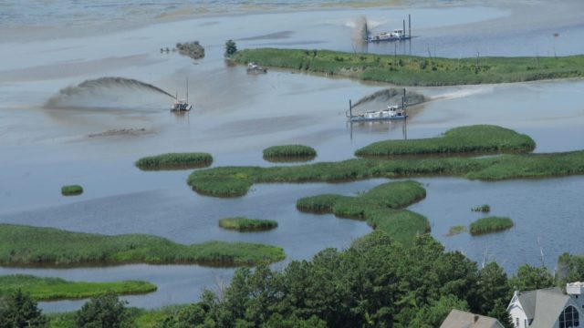 Aerial view of a coastal marsh restoration site where several floating dredging units spray sediment slurry across shallow water and fragmented marsh, surrounded by patches of dense green vegetation.