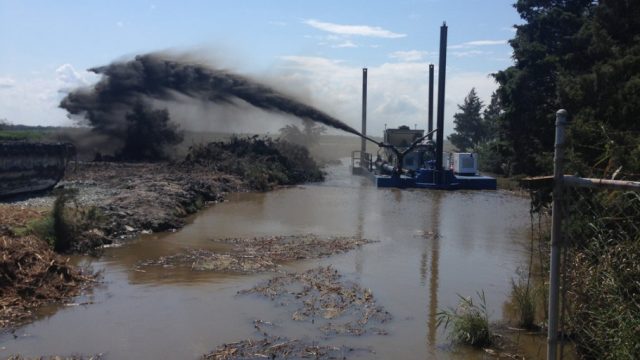 Dredging or sediment-placement equipment pumps a dark slurry through a pipe across a flooded wetland restoration site, with the material spraying onto the adjacent marsh surface.