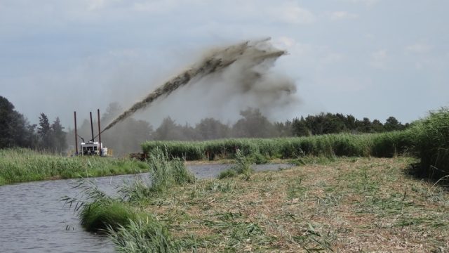 Sediment-placement equipment sprays a high arc of dark slurry across a marsh restoration site, distributing material over dense wetland vegetation beside a narrow water channel.