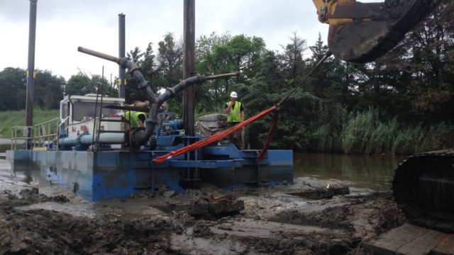 Construction crew operating a barge-mounted pumping or dredging system in a muddy wetland area. An excavator bucket and rigging are visible in the foreground, while workers in high-visibility safety gear stand on the equipment beside a narrow waterway.